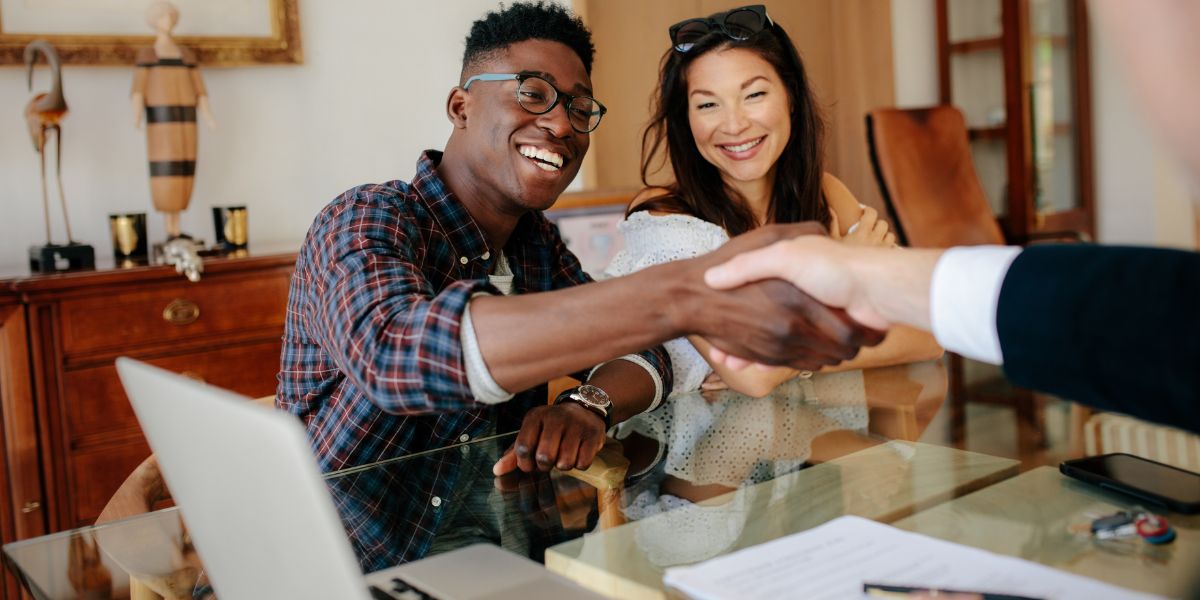 Smiling couple shaking hands with a real estate agent across a glass table.