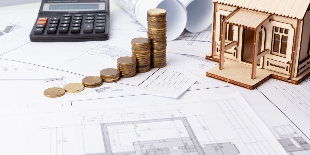 Coins stacked on architectural blueprints next to a calculator and a wooden house model.