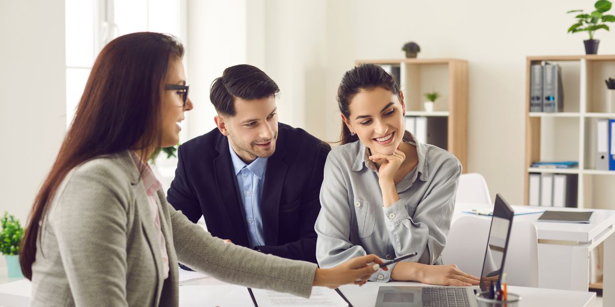 Real estate agent showing information on a laptop to a smiling couple in an office setting.