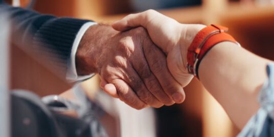 Professional buyer's agent and listing agent shaking hands across a modern wooden table, symbolizing a successful real est...